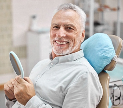 An older man wearing a button-down shirt and holding a handheld mirror while smiling and showing off his new dentures in Hillsboro